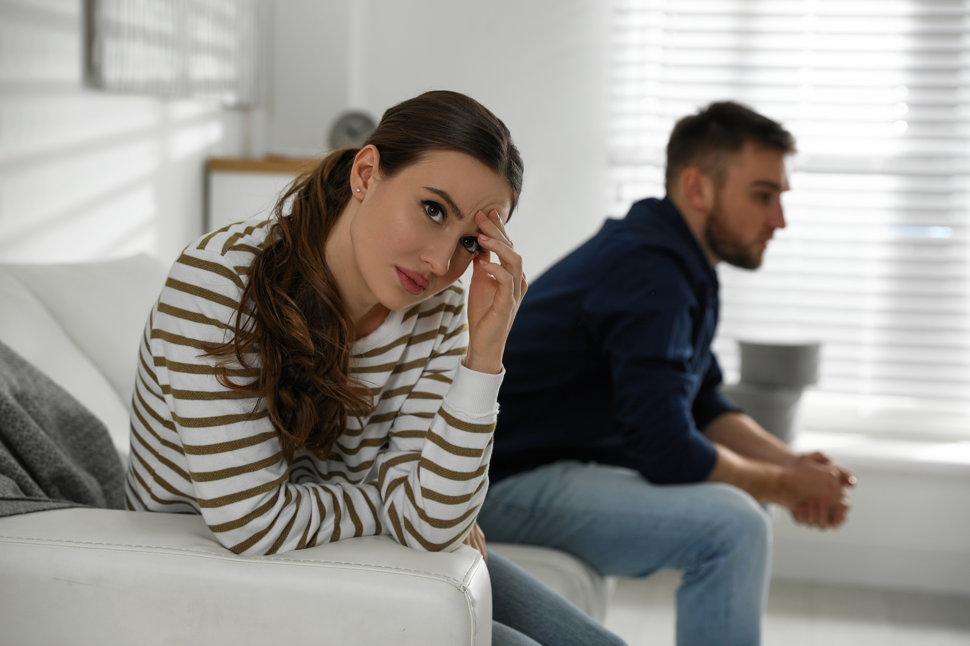 A woman with long brown hair, wearing a striped sweater, sits on a white couch looking concerned. In the background, a man with short hair, wearing a navy hoodie and jeans, sits on the edge of the couch facing away, both appearing upset in a bright room.