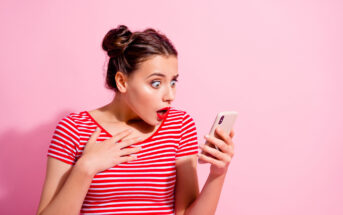 A young woman with two buns in her hair and wearing a red and white striped shirt is looking at her phone with a surprised expression. Her eyes are wide open, and her mouth is slightly agape. She is against a solid pink background.