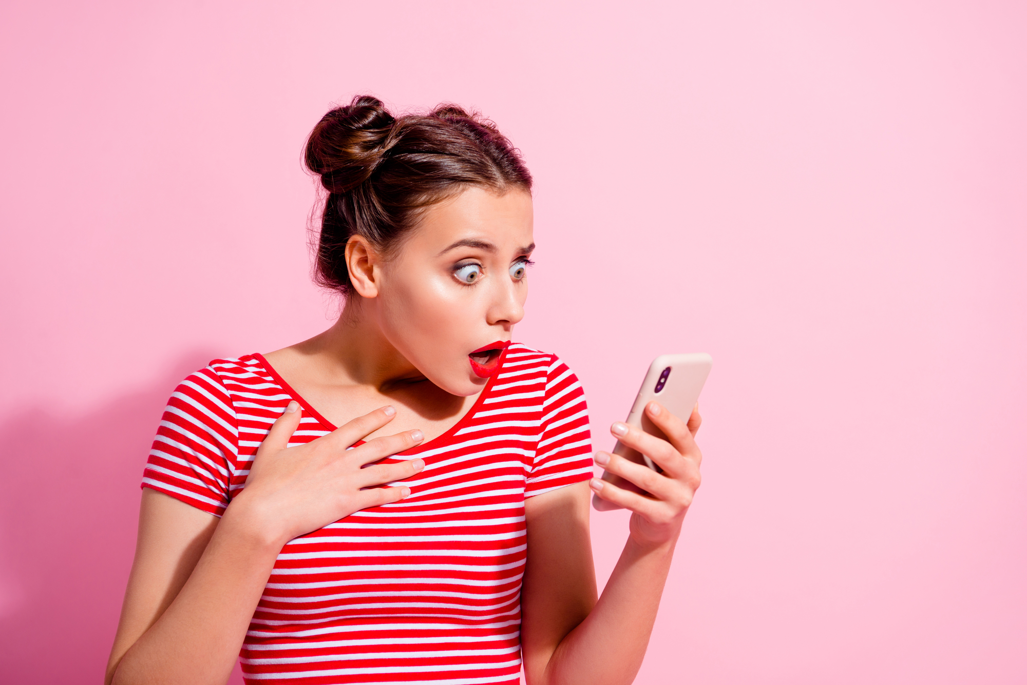 A young woman with two buns in her hair and wearing a red and white striped shirt is looking at her phone with a surprised expression. Her eyes are wide open, and her mouth is slightly agape. She is against a solid pink background.