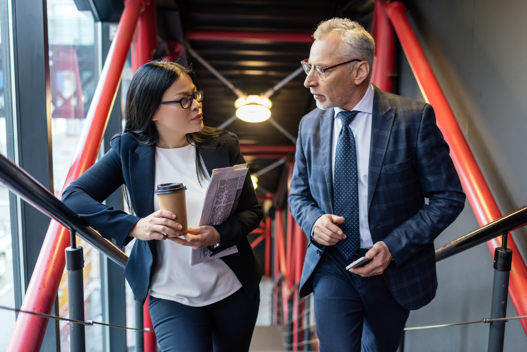 Two business professionals walking and talking. A woman with glasses carries a coffee cup and a notebook, while a man with glasses dressed in a suit holds a smartphone. They are walking through a modern hallway with red beams and glass walls.