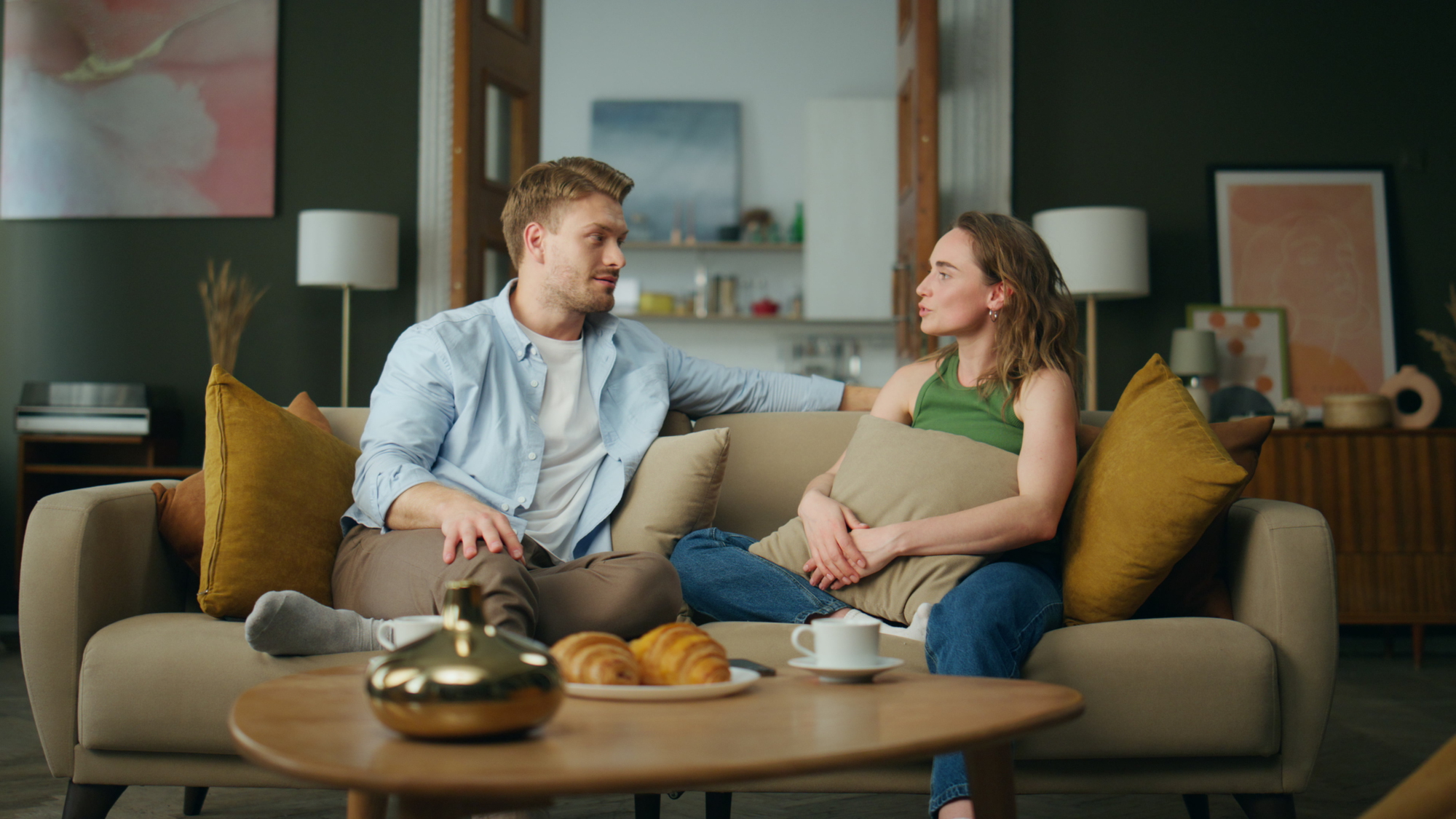 A man and woman sit closely together on a beige sofa in a cozy living room. The man has his arm around the back of the sofa, while the woman holds a pillow and looks at him. A wooden coffee table in front of them has croissants and two cups of coffee.