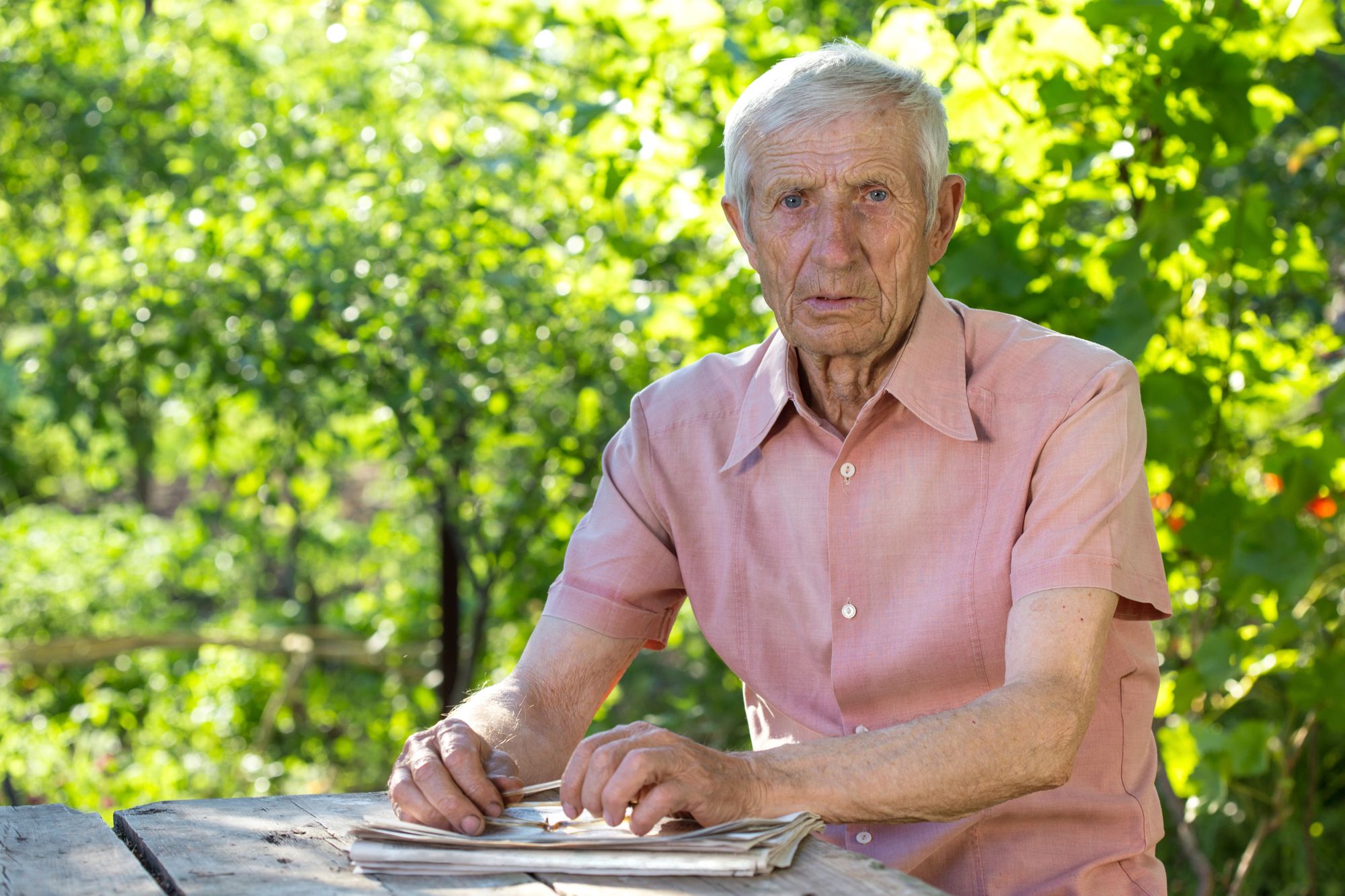 An elderly man with short white hair, wearing a pink short-sleeved button-up shirt, sits at a wooden table outside with greenery in the background. He looks directly at the camera, his hands resting on a stack of newspapers on the table.