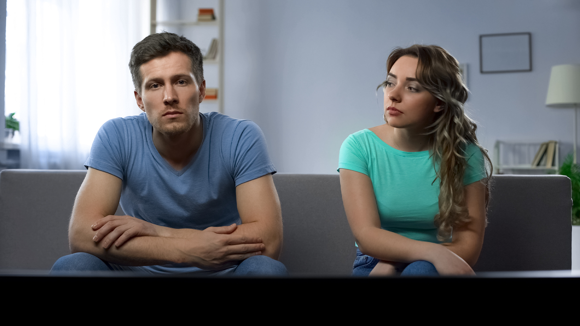 A man and a woman sit on a couch in a living room, both looking towards the camera with serious expressions. The man wears a blue t-shirt and the woman wears a teal t-shirt. The room has white walls with shelves, a lamp, and a framed picture in the background.