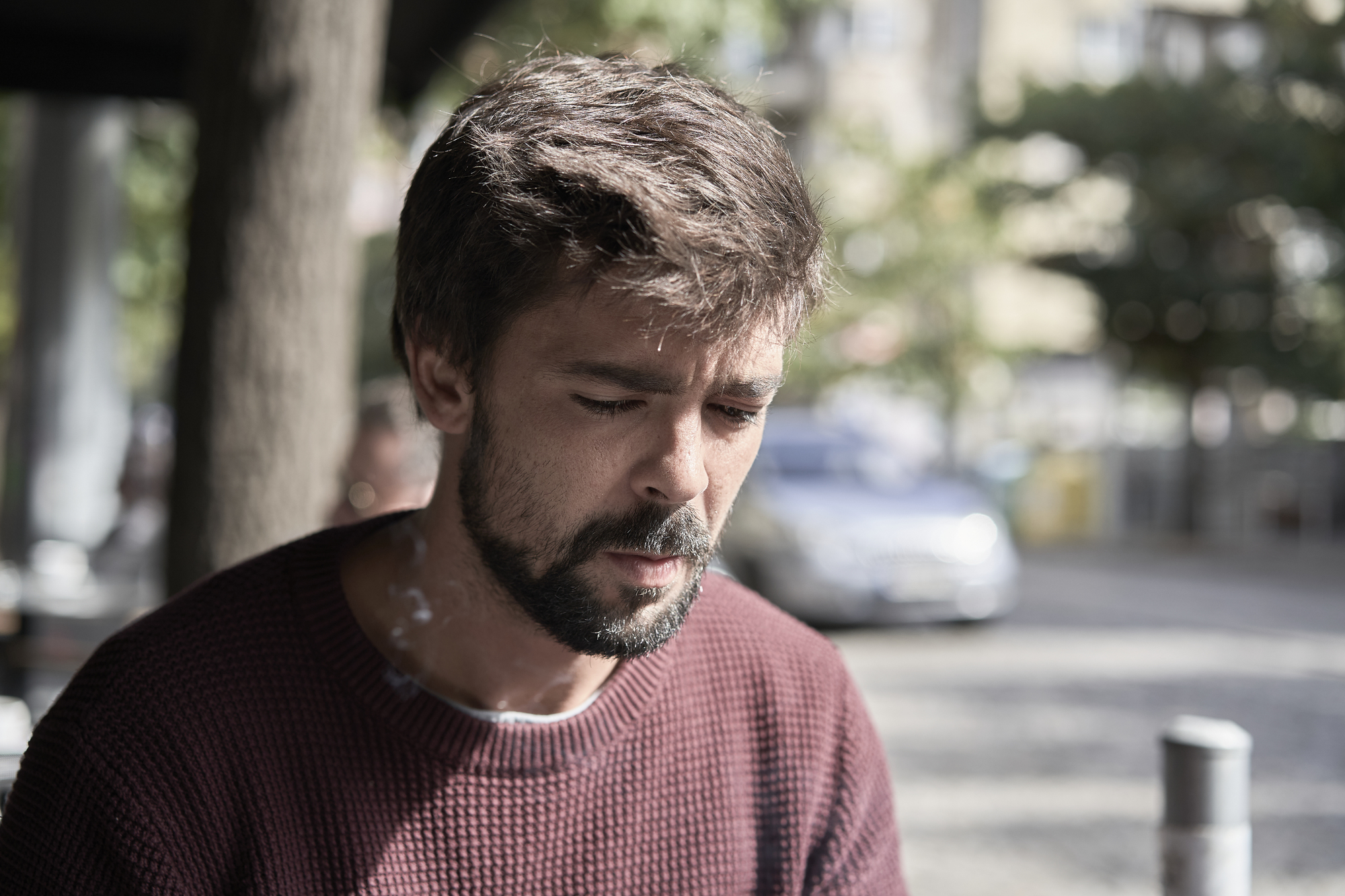 A man with a beard and mustache, wearing a maroon sweater, is looking down pensively. He is outdoors on a sunny day, with blurred trees and a parked car in the background. Smoke or mist is visible around his face.