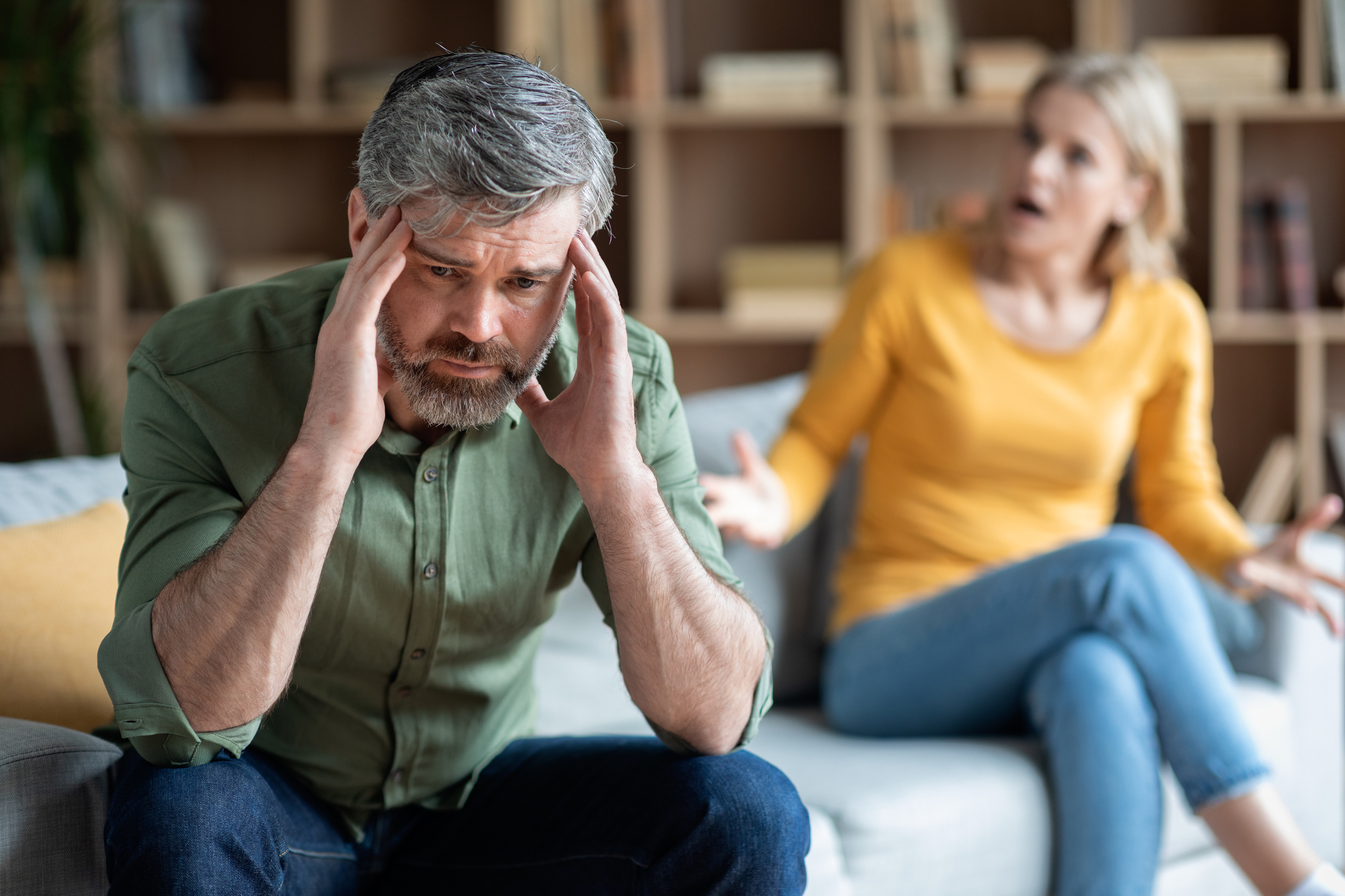 A man with gray hair and beard, wearing a green shirt, sits on a couch holding his head in his hands, looking distressed. In the background, a woman with blonde hair, wearing a yellow shirt, appears to be arguing or talking animatedly.