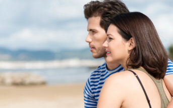 A man and a woman are standing close together, facing the same direction and looking at the horizon. They appear to be on a beach with sand and water in the background. Both are casually dressed and have a calm expression on their faces under a cloudy sky.