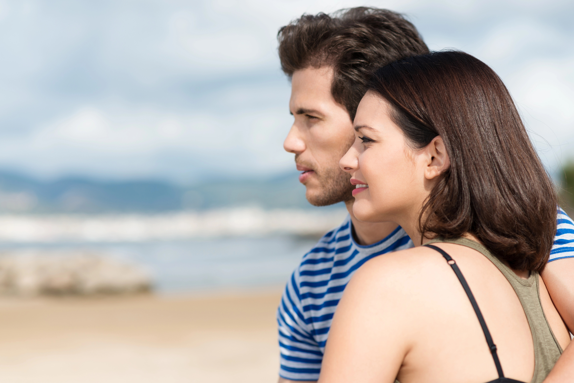 A man and a woman are standing close together, facing the same direction and looking at the horizon. They appear to be on a beach with sand and water in the background. Both are casually dressed and have a calm expression on their faces under a cloudy sky.