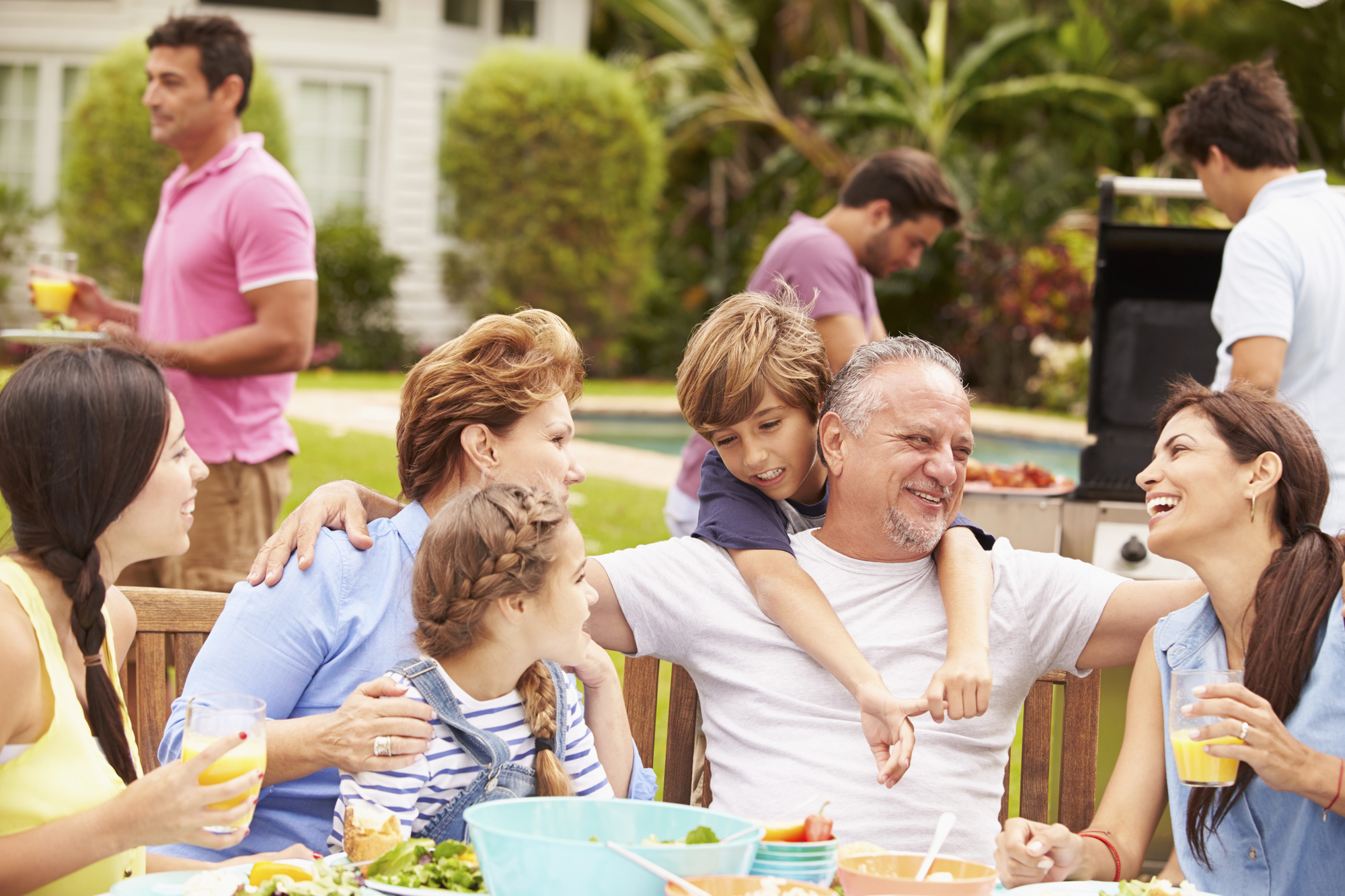 A multigenerational family gathers outdoors around a table, sharing a meal and laughing. A young boy hugs an older man, while others share smiles and conversation. People in the background are preparing food on a grill. Lush greenery and a house are visible.