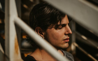 A person with medium-length dark hair is seen in close-up, standing behind a white metal railing. Their eyes are closed, and they appear to be in deep thought or relaxation, with soft sunlight casting shadows on their face.