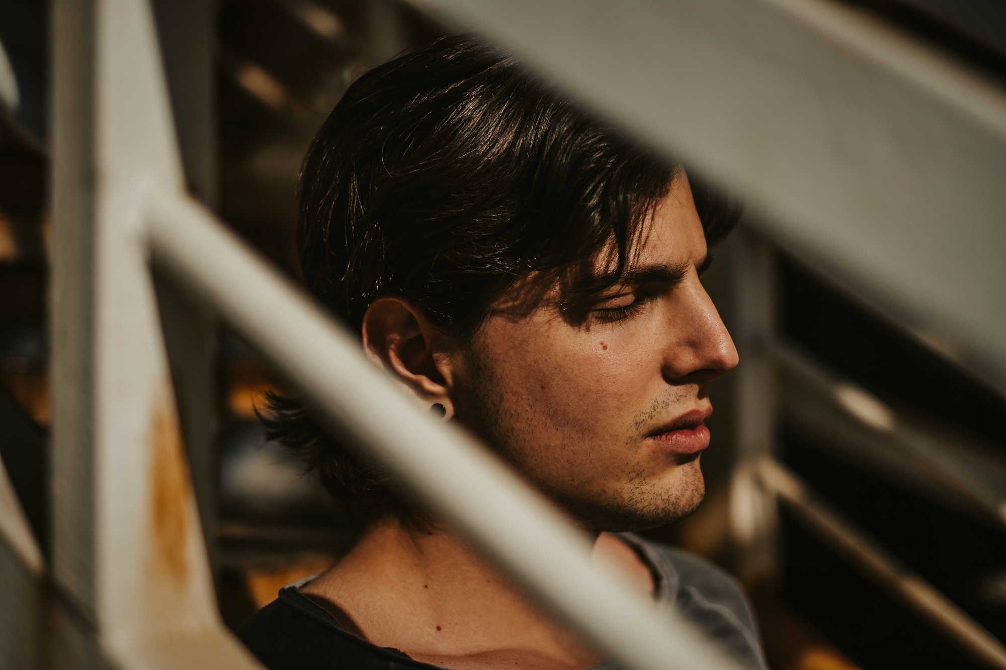 A person with medium-length dark hair is seen in close-up, standing behind a white metal railing. Their eyes are closed, and they appear to be in deep thought or relaxation, with soft sunlight casting shadows on their face.