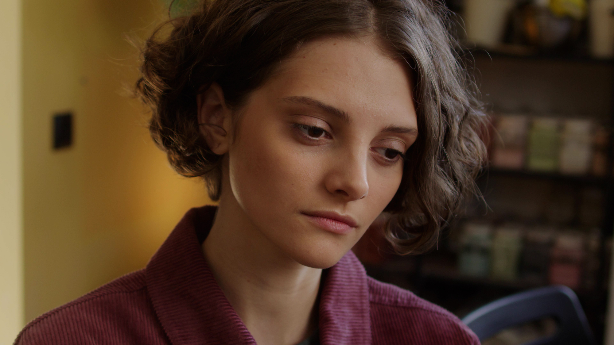 A young woman with short, wavy brown hair and a maroon collared shirt gazes downward with a thoughtful expression. The background is softly blurred, featuring warm colors and indistinct objects, suggesting an indoor setting.