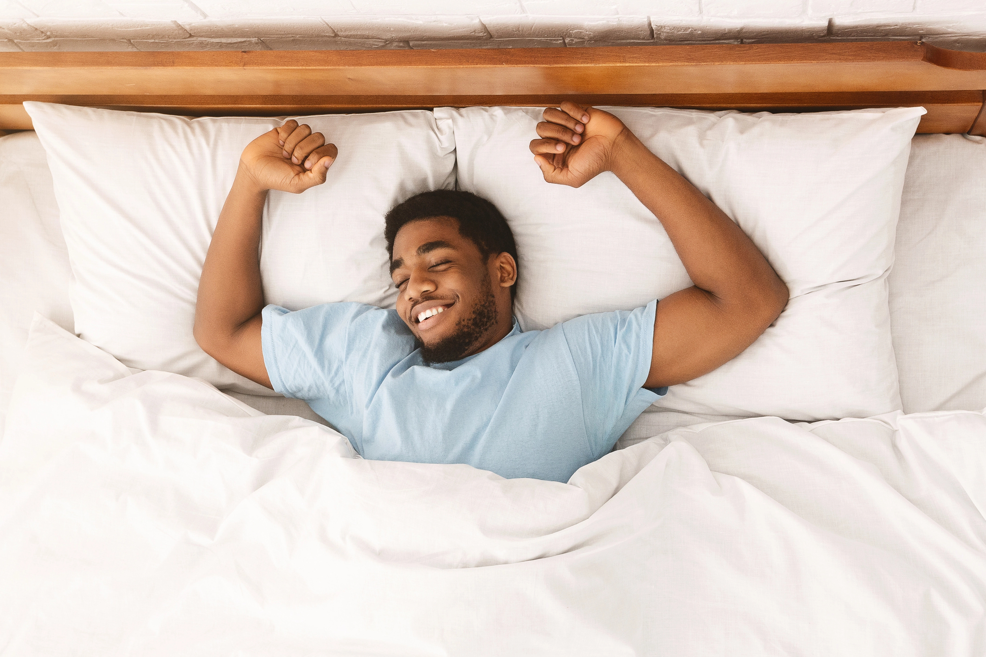 A man wearing a light blue shirt is lying in bed, smiling, and stretching with both arms raised above his head. He is resting on white pillows and covered with a white blanket. The headboard is wooden.