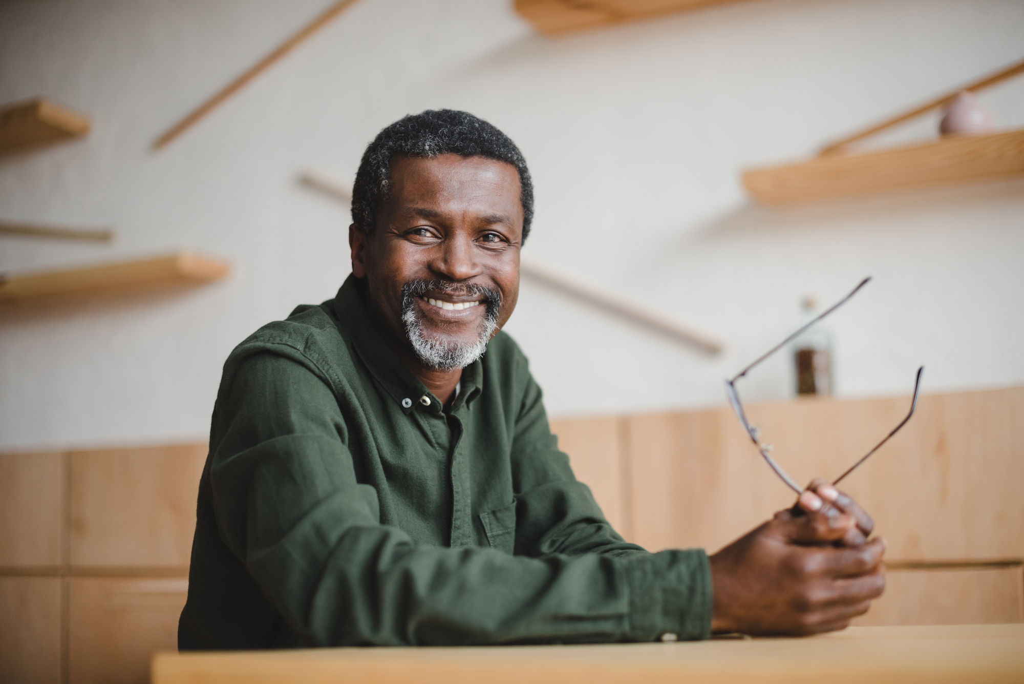 A man with short curly hair and a beard, wearing a dark green shirt, is sitting at a wooden table indoors. He is smiling at the camera and holding a pair of glasses in his hands. The background features wooden shelves mounted on a light-colored wall.