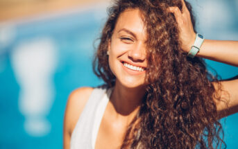 A woman with long, curly hair smiles brightly with one hand in her hair. She is wearing a white sleeveless top and a wristband, and is standing in front of a blue, blurry background, possibly water.