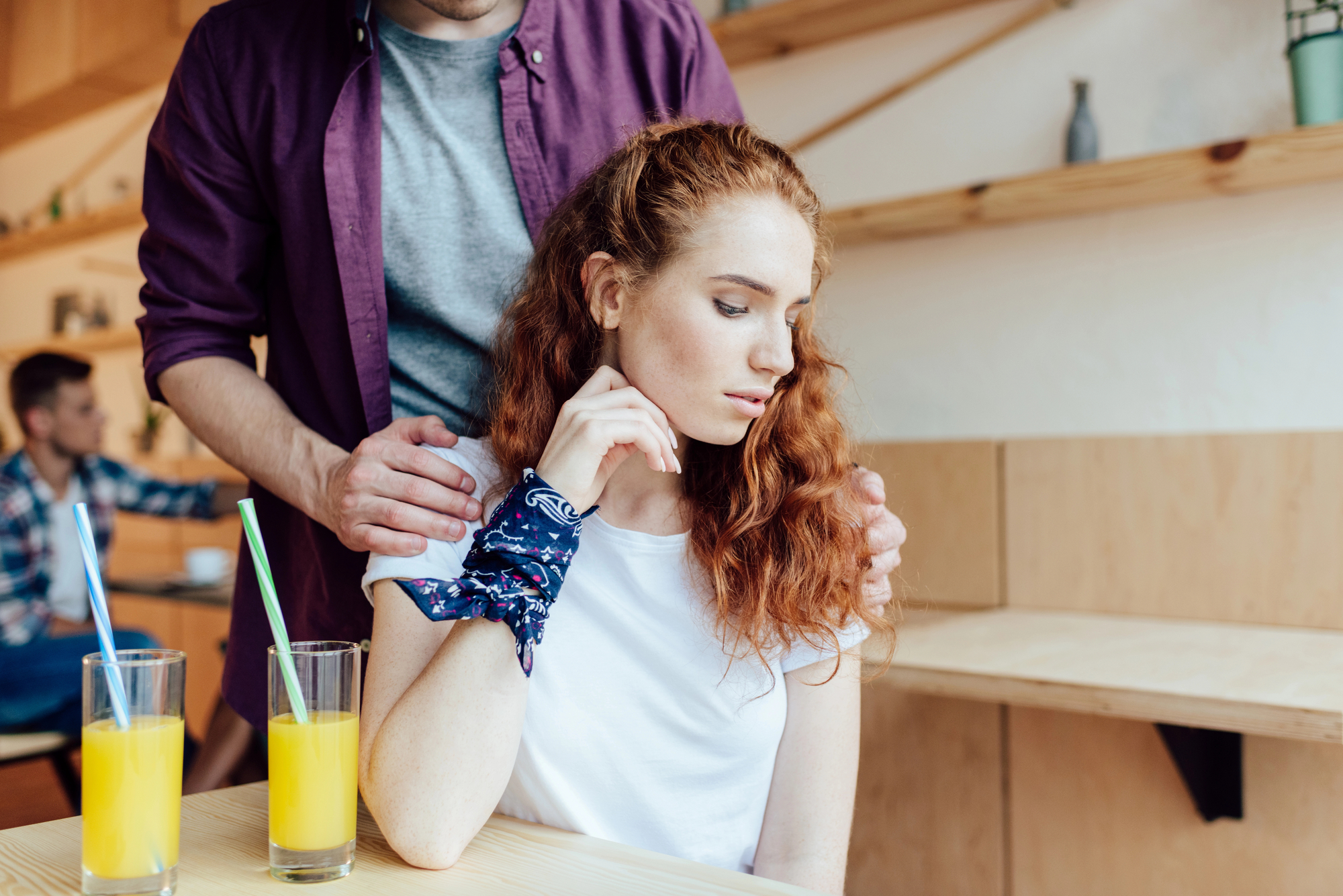 A woman with red curly hair sits at a table looking thoughtful, resting her head on her hand. A man stands behind her with his hands gently on her shoulders. Two glasses of orange juice with straws are in the foreground. They are in a casual, modern café.