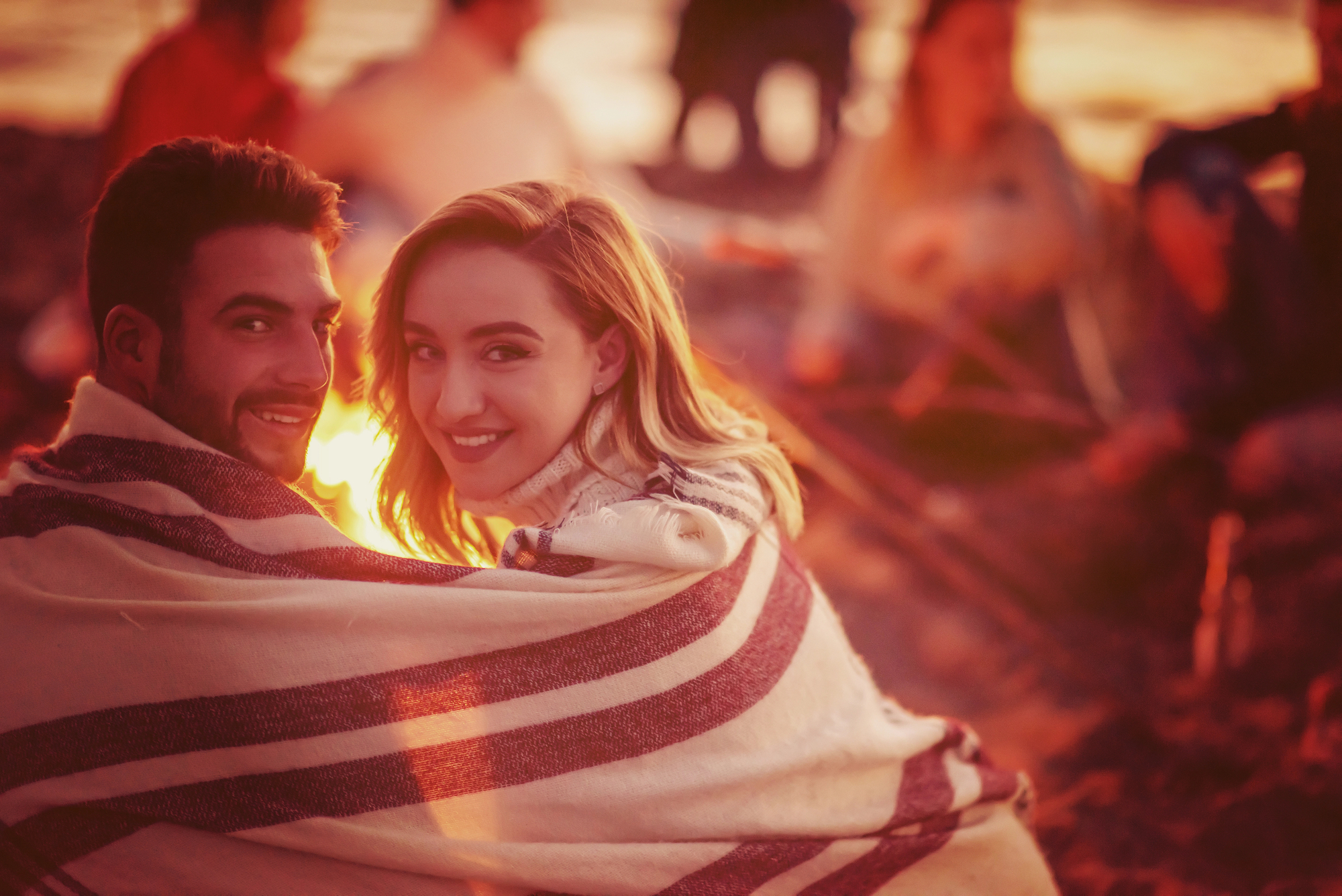 A couple sits on a beach wrapped in a striped blanket, smiling at the camera as the sun sets. Behind them, friends are gathered around a campfire, enjoying the warm glow and relaxed atmosphere. The scene is lit with golden, ambient light, evoking a cozy, intimate feel.