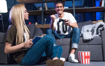 A young man is sitting on a couch and playing video games with a game controller, smiling enthusiastically. A young woman is seated on the floor in front of him, reading a book. A striped popcorn container is on the floor beside them. The room has shelves and soft lighting.