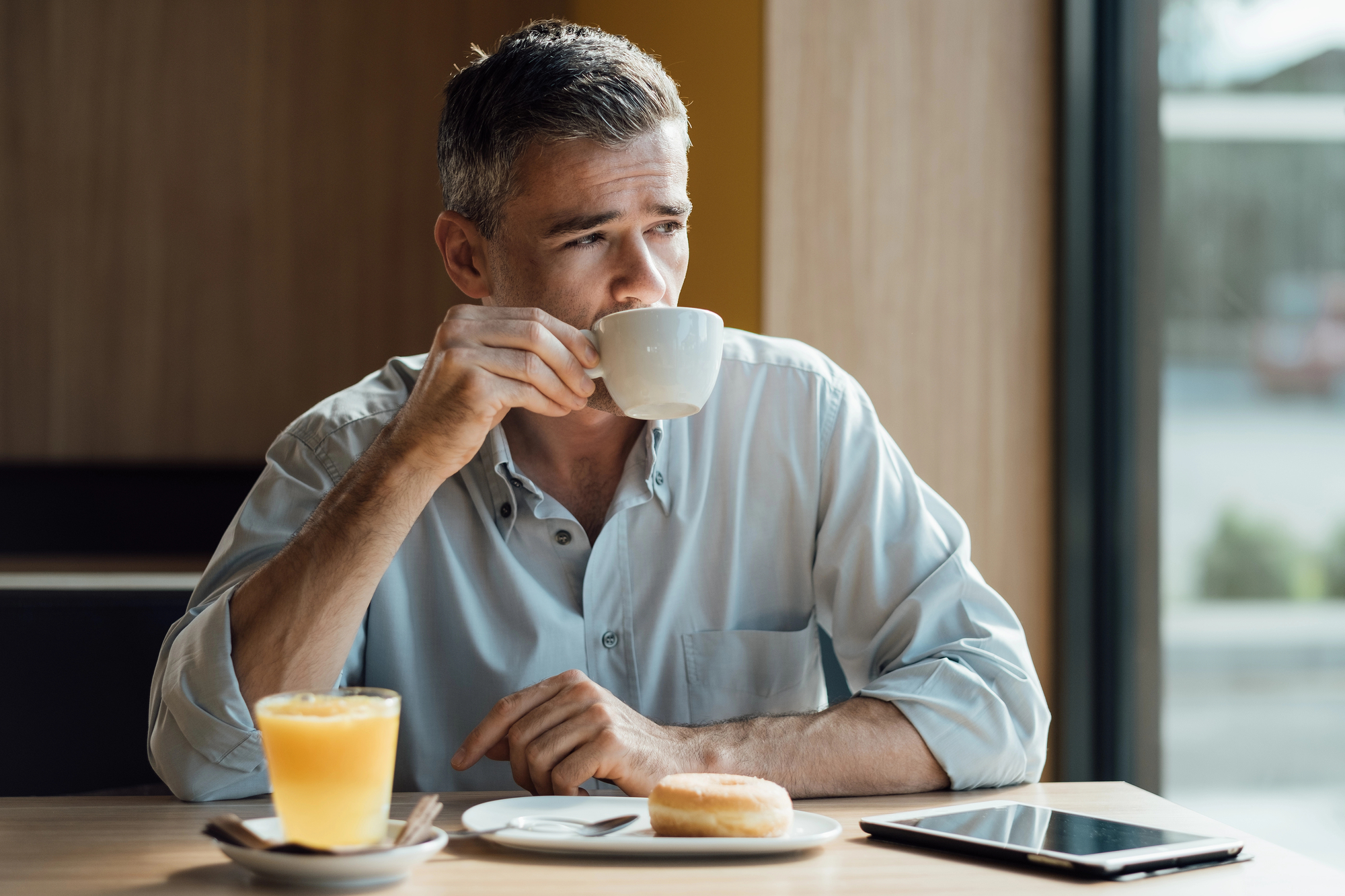 A man with salt-and-pepper hair sips from a white coffee cup while sitting at a table in a café. On the table are a glass of orange juice, a donut on a plate, and a tablet. He gazes out of the window, appearing thoughtful.