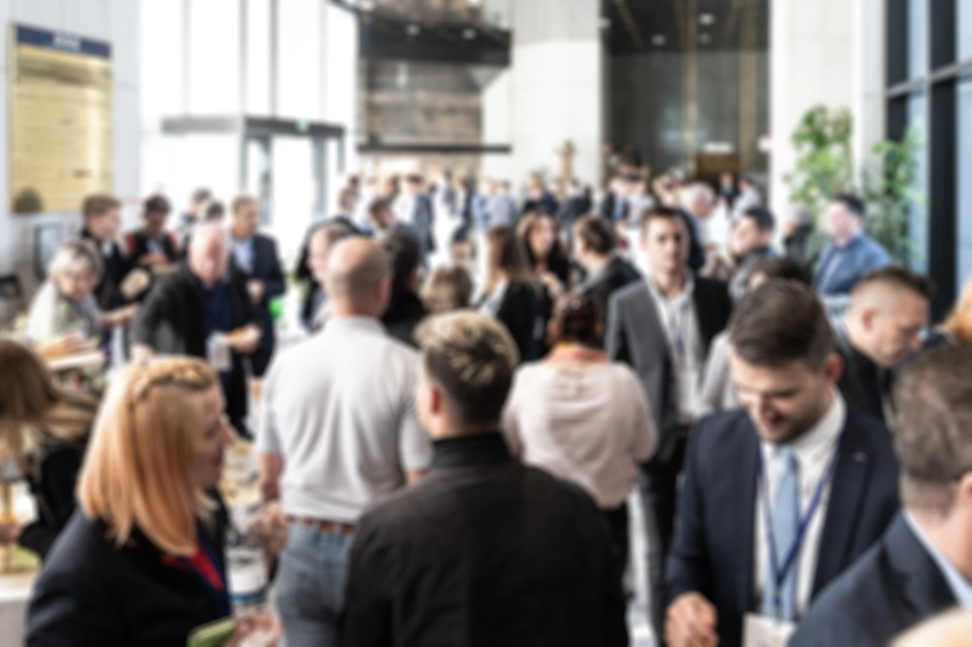 A blurred image showing a large group of people gathered in what appears to be an indoor event or conference. The individuals are engaged in conversations, with some holding drinks and papers. The setting includes large windows and modern architecture.