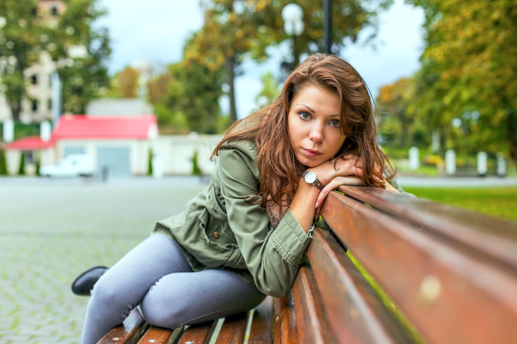 A young woman with long brown hair and a green jacket leans on a wooden bench outdoors, looking directly at the camera. Trees and buildings are visible in the background, suggesting a park or urban setting. The expression on her face is pensive.