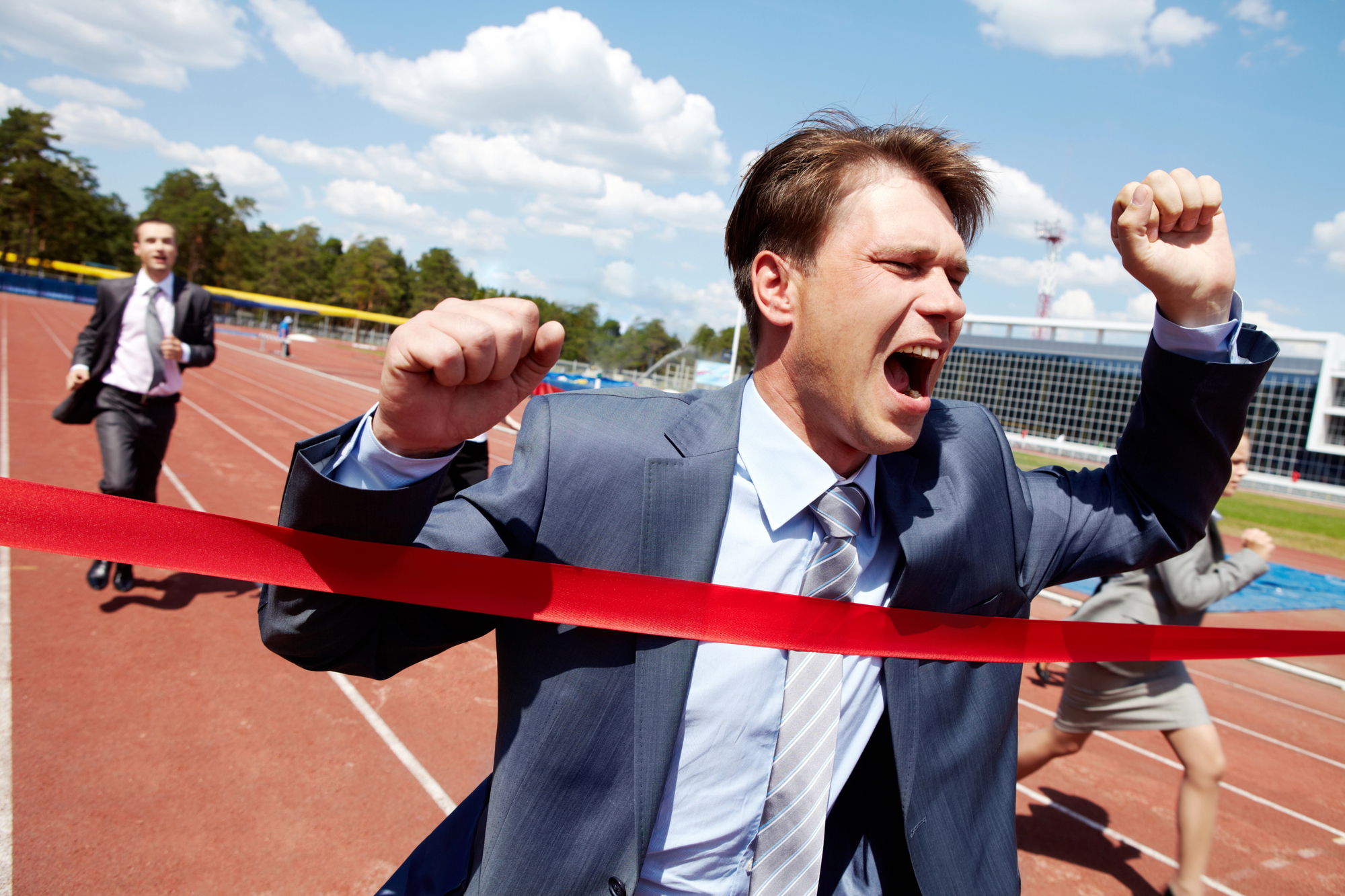 A man in a suit energetically crosses a red finish line on a running track, appearing triumphant with his arms raised and mouth open in a victory shout. Other individuals in business attire are running behind him. The background shows trees, buildings, and a partly cloudy sky.