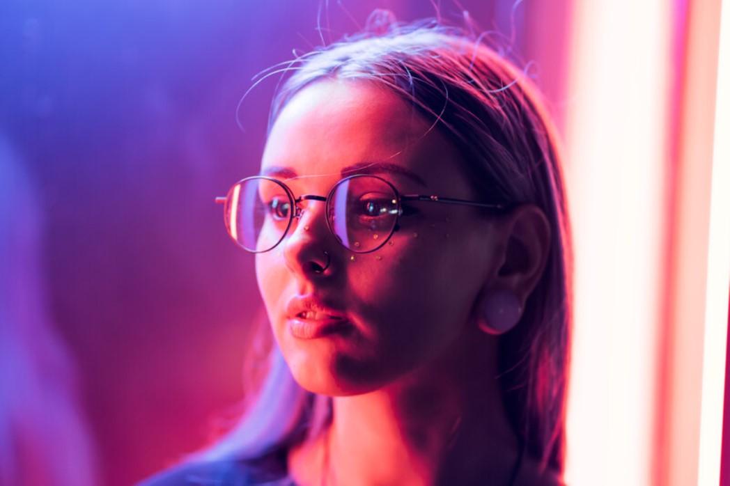 A young woman with glasses and a nose piercing looks to the side, illuminated by vibrant pink and purple neon lights. She has long hair and large ear gauges, creating an atmospheric and contemporary scene.