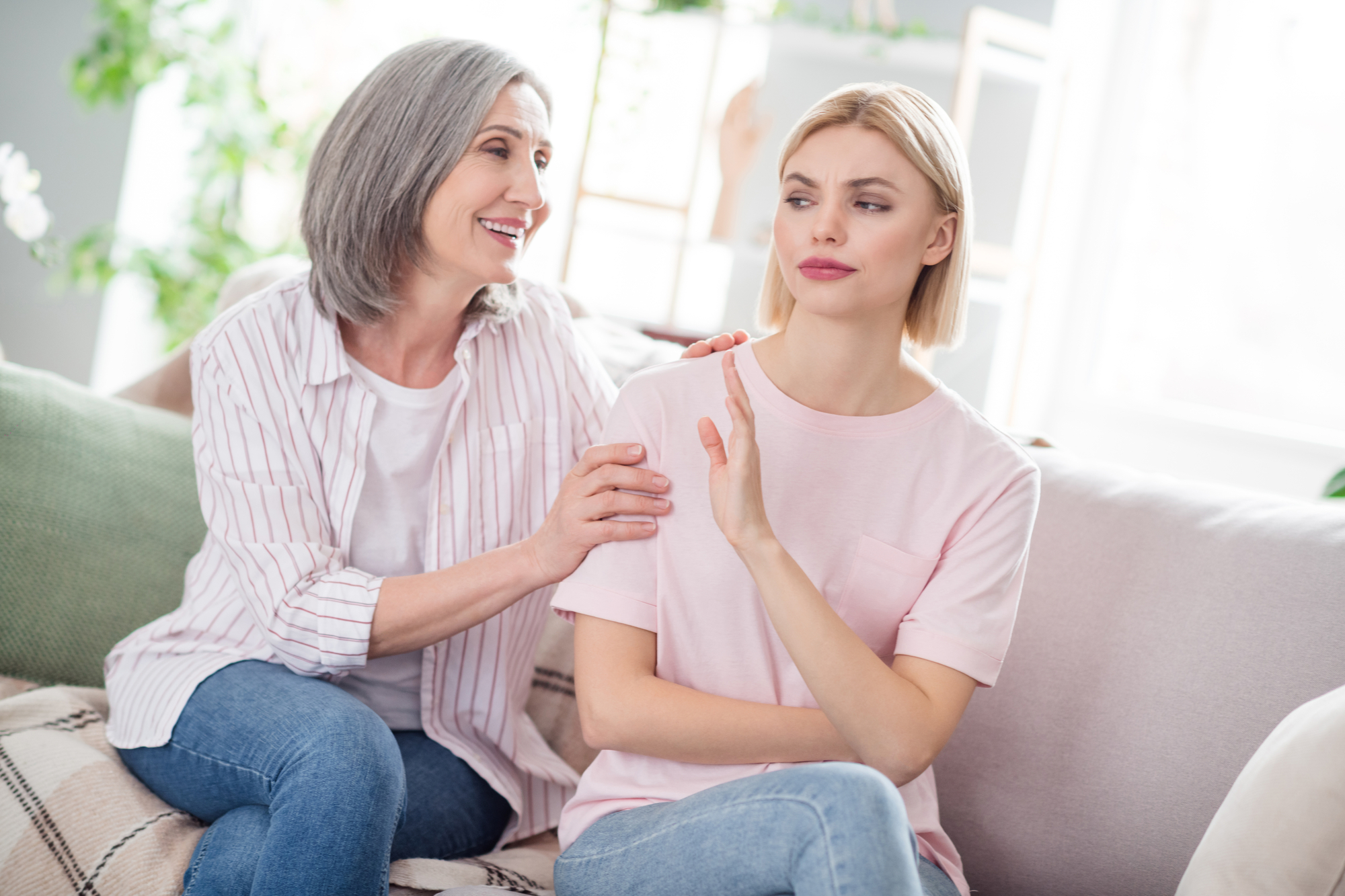 A middle-aged woman with gray hair, wearing a striped shirt, sits next to a younger woman with blonde hair, in a pink shirt, on a couch. The older woman smiles and touches the younger woman's shoulder, who looks away with a hand raised in a dismissive gesture.