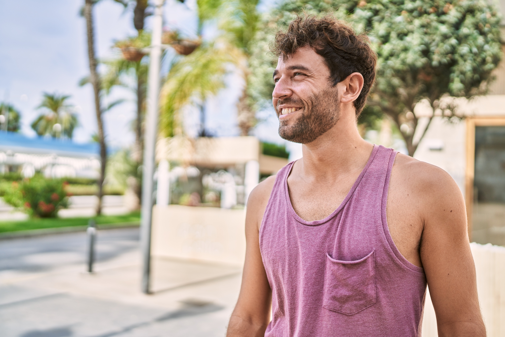 A man with curly hair and a beard smiles while standing outdoors. He is wearing a sleeveless purple top with a pocket, and palm trees are visible in the background. The scene appears to be in a sunny, urban area with a relaxed atmosphere.