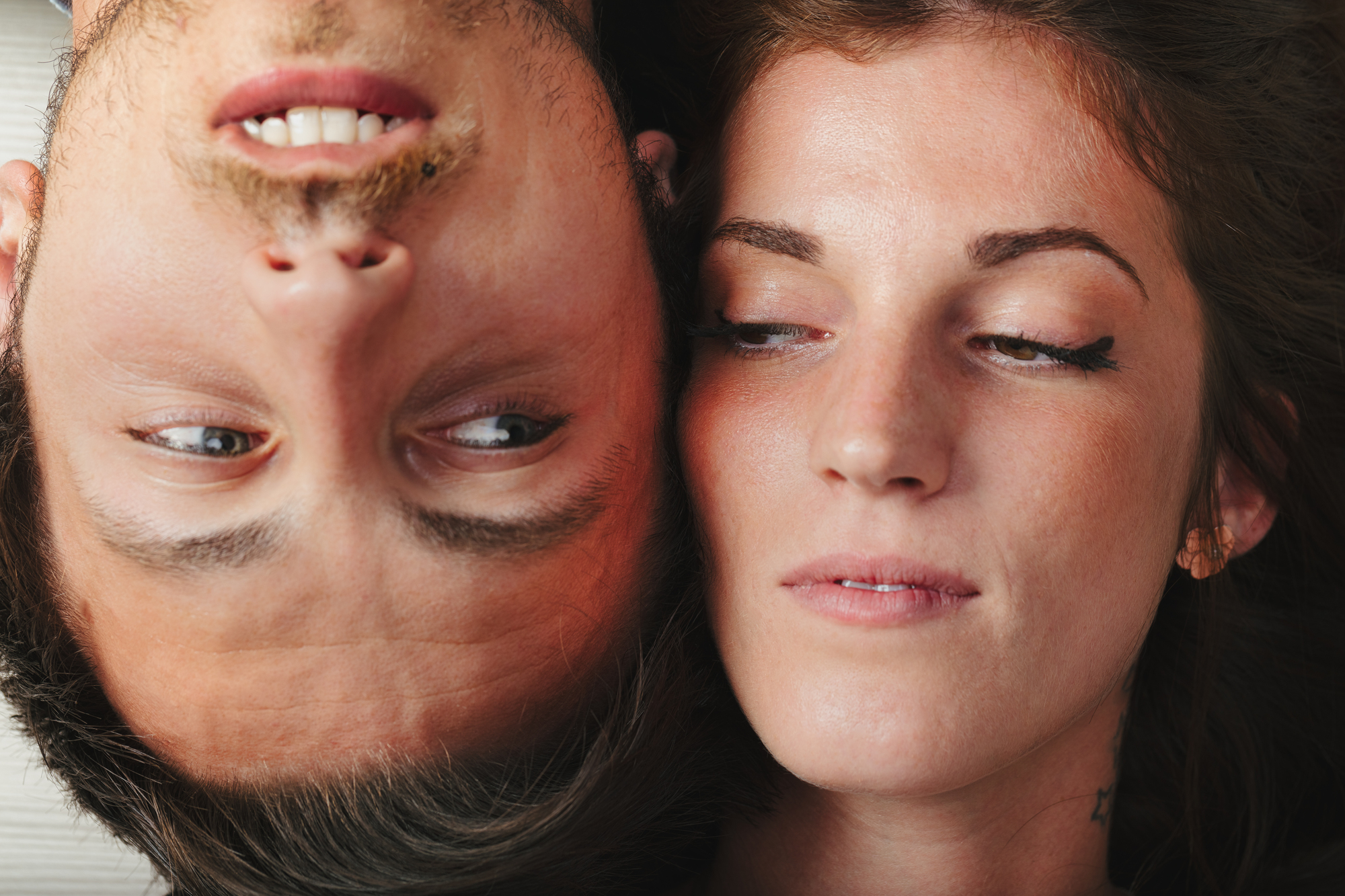 Close-up of two people lying down, facing each other closely. One person's face is upside down, and they have a slight smile. The other person's head is right-side up, and they have a neutral, slightly inquisitive expression. They both have light complexions.