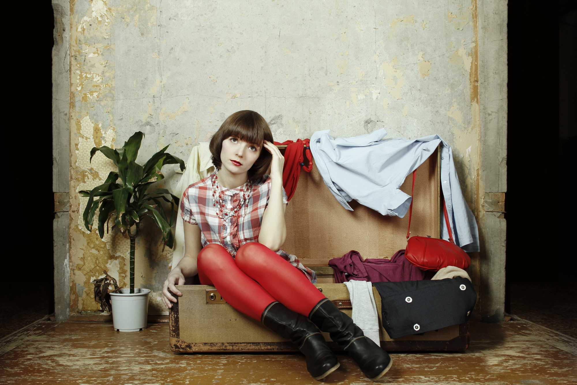 A woman with short brown hair sits inside an open suitcase placed against a worn wall. She wears a plaid shirt, red tights, and black boots. Clothes are strewn around her. A potted plant is on her left, and she rests her head on her hand with a thoughtful expression.