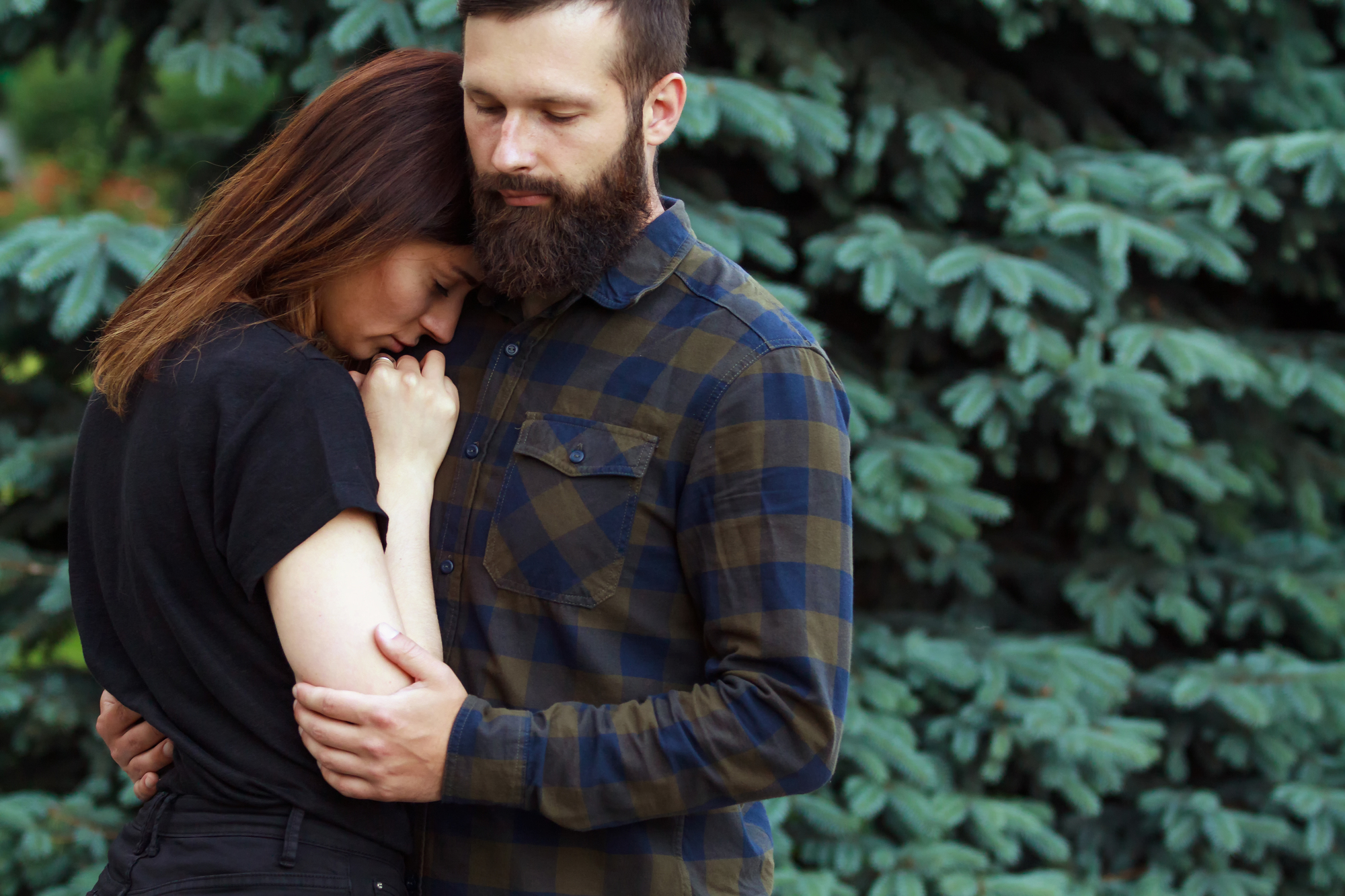 A bearded man wearing a plaid shirt embraces a woman with long, brown hair, who is leaning against his chest with a somber expression. They are standing in front of a large pine tree. The scene suggests comfort and support.