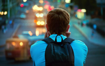 A person with short hair wearing a blue shirt and a black backpack is seen from behind, gazing at a city street filled with blurred traffic lights and vehicles during dusk. The scene conveys a sense of contemplation and urban life.