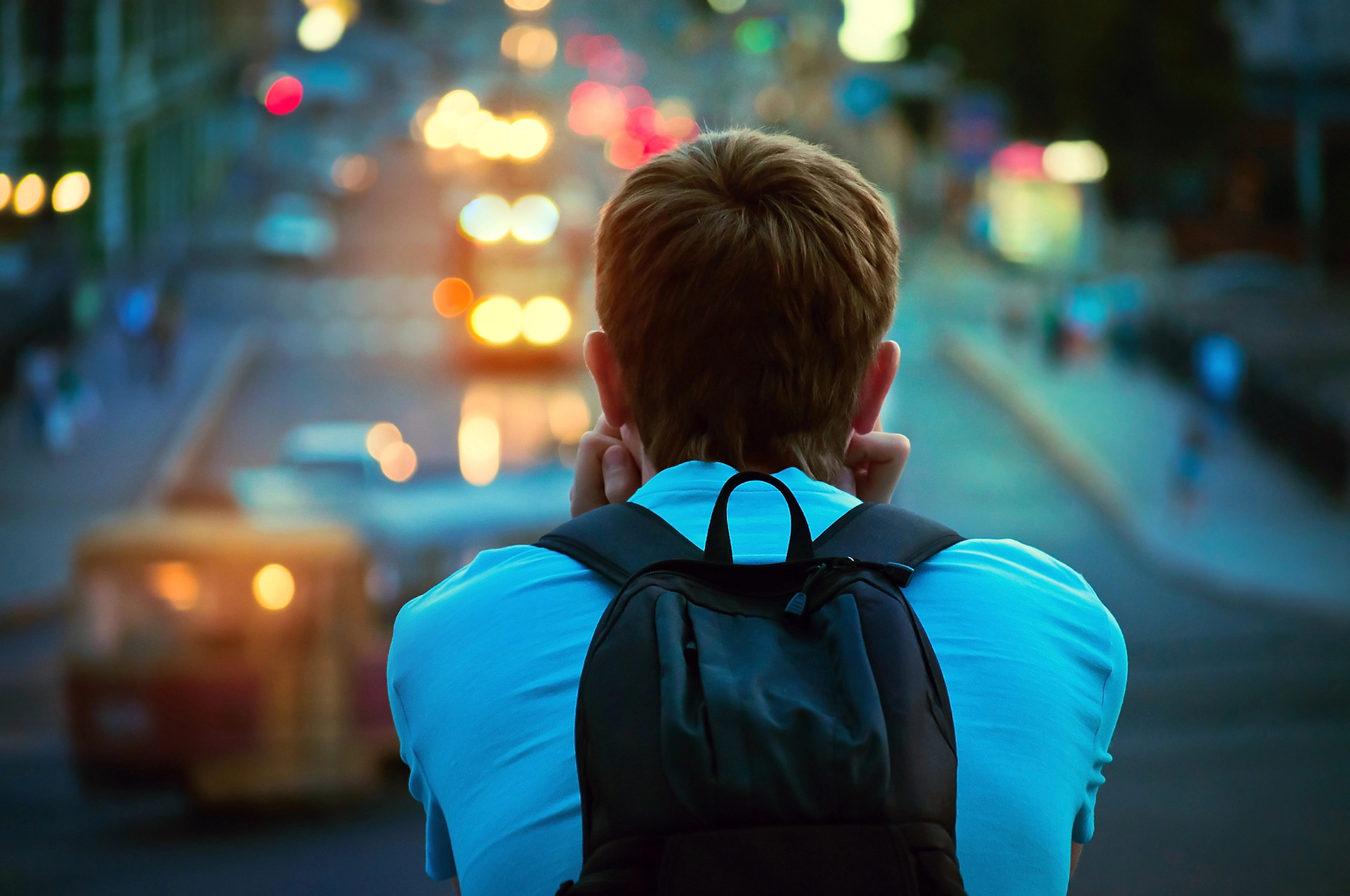 A person with short hair wearing a blue shirt and a black backpack is seen from behind, gazing at a city street filled with blurred traffic lights and vehicles during dusk. The scene conveys a sense of contemplation and urban life.