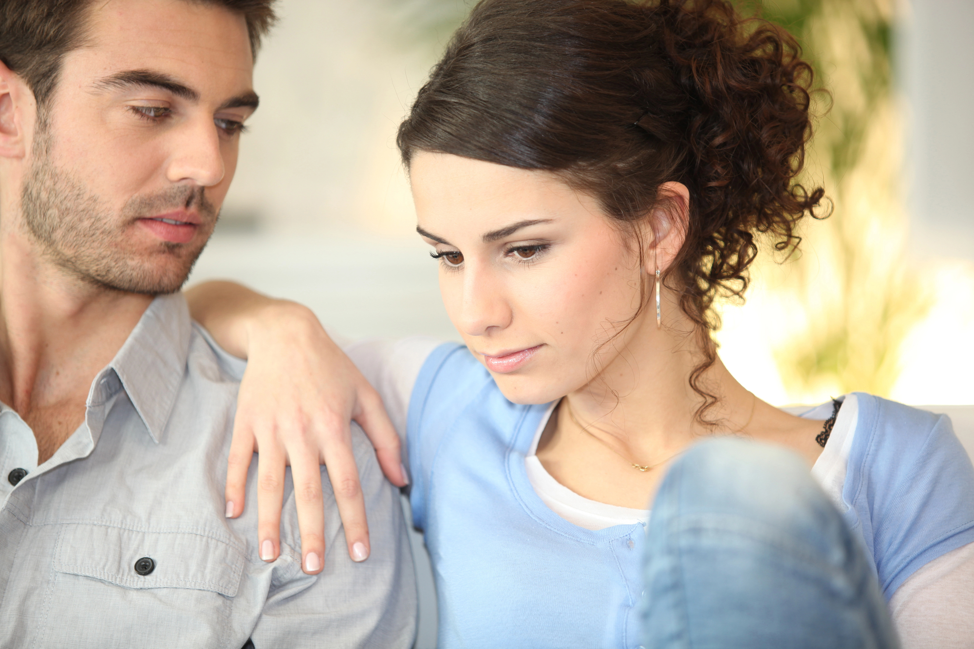 A man and a woman sit closely together, with the man gently resting his arm around the woman's shoulder. The woman appears thoughtful, looking downwards, while the man looks at her with concern. Both are casually dressed and positioned in a softly lit environment.