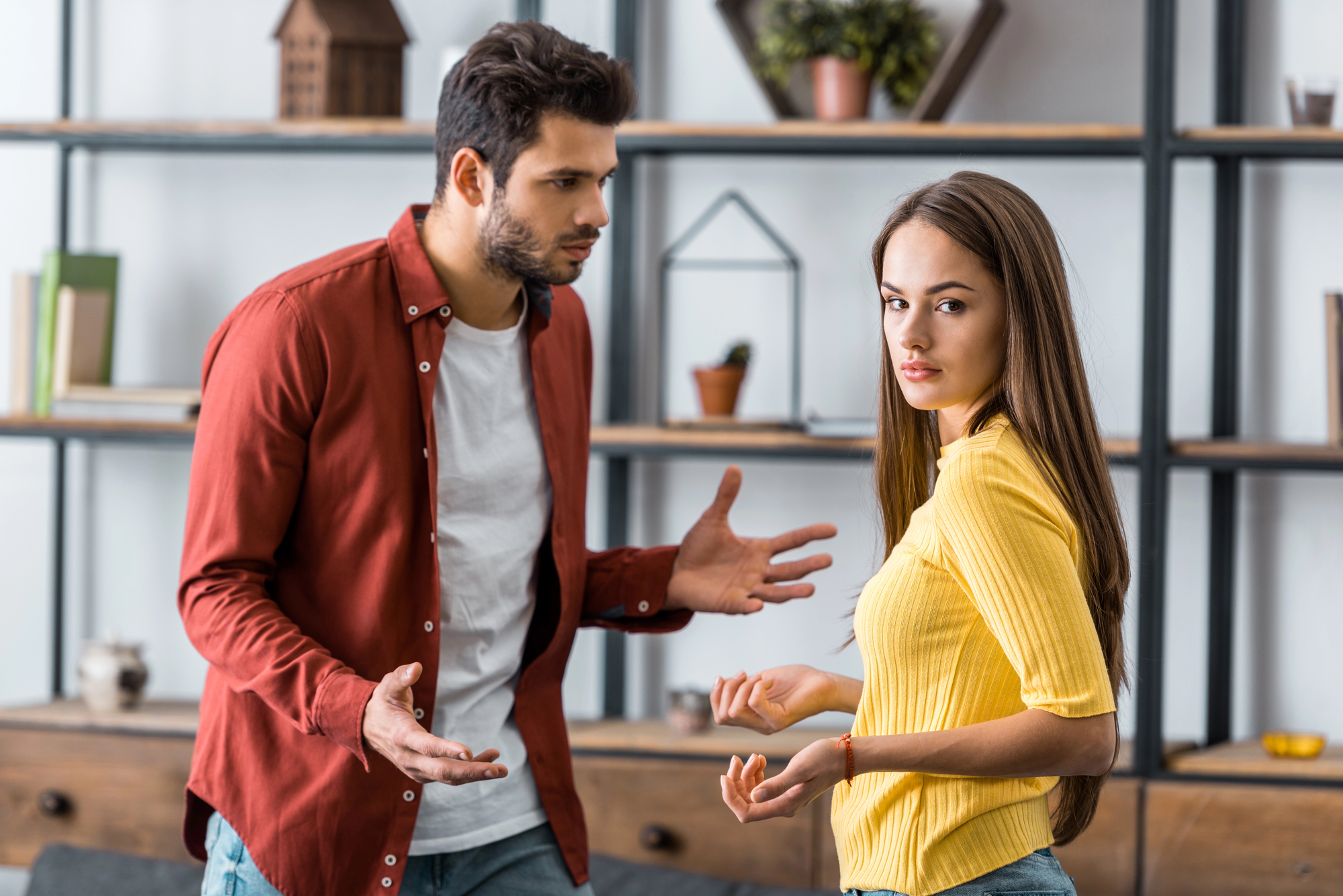 A man and a woman are having a tense conversation indoors. The man, wearing a red shirt, gestures with his hands while speaking, and the woman, in a yellow sweater, looks away with a troubled expression. They are standing in front of shelves with various decorations.