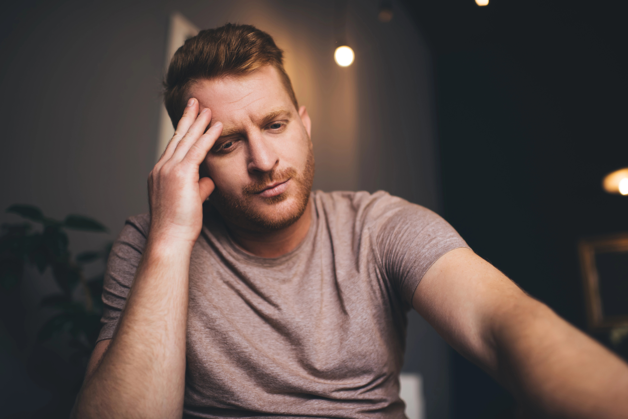 A man with a short, brown hair and a beard, wearing a light purple t-shirt, sits indoors with a pensive expression, resting his head on his hand. The background is dimly lit with a warm light bulb visible.