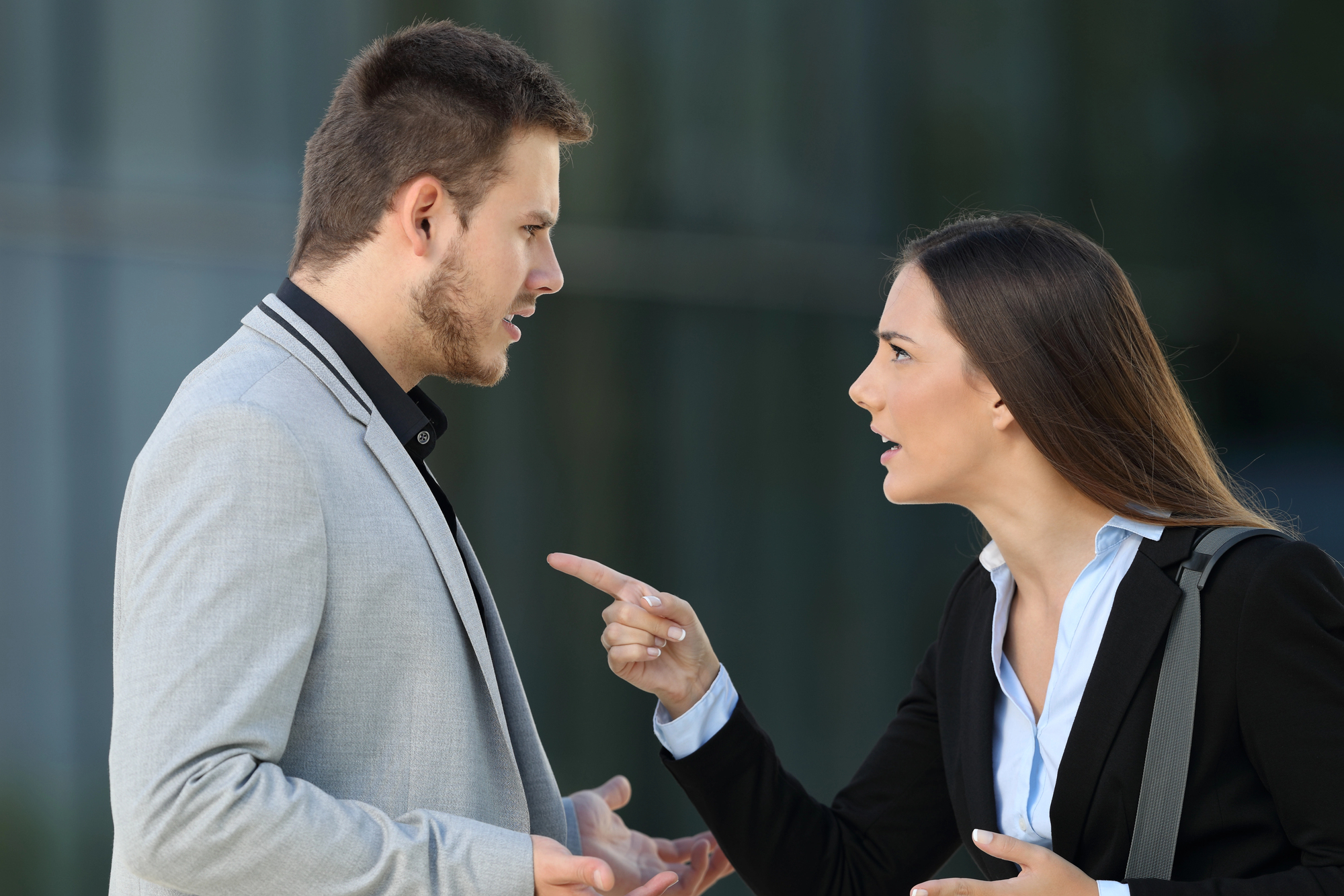 A man and a woman are having a heated argument outdoors. The woman, wearing a black blazer, is pointing her finger and appears to be speaking angrily. The man, dressed in a grey blazer, looks confused and is gesturing with his hands.