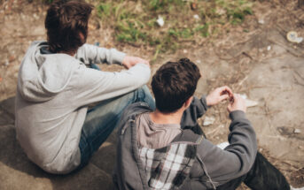 Two young people sitting on outdoor steps. They both wear hooded jackets, and are seated next to each other facing slightly away from the camera. One has dark hair, while the other has brown, and they appear to be engaged in conversation. A grassy area is in the background.