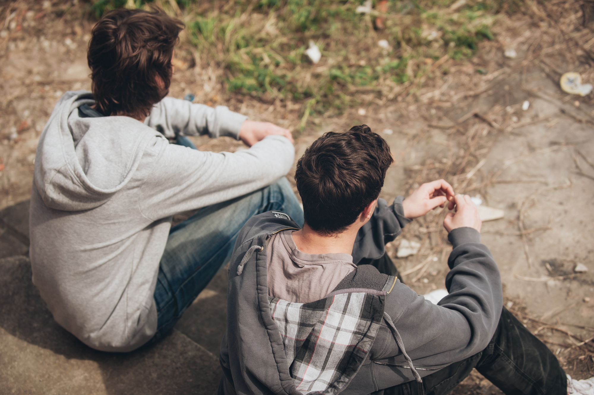 Two young people sitting on outdoor steps. They both wear hooded jackets, and are seated next to each other facing slightly away from the camera. One has dark hair, while the other has brown, and they appear to be engaged in conversation. A grassy area is in the background.