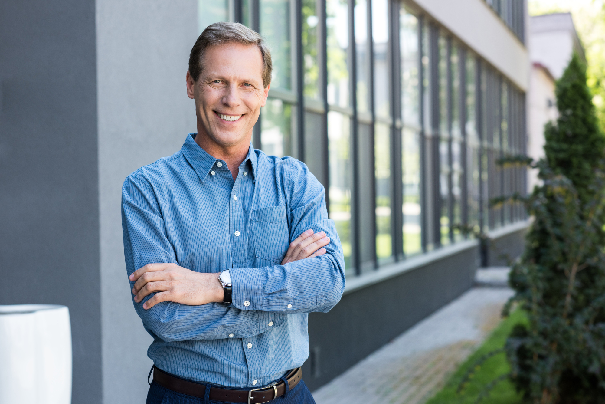 A man wearing a blue button-up shirt stands outside a modern building with large windows, smiling with his arms crossed. The background includes green foliage along the building's exterior. The scene is bright and appears to be a sunny day.