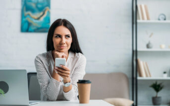 A woman with long dark hair sits at a desk holding a smartphone and smiling thoughtfully. She is in a modern, well-lit room with a laptop in front of her, a coffee cup next to her, a bookshelf in the background, and a painting on the wall.