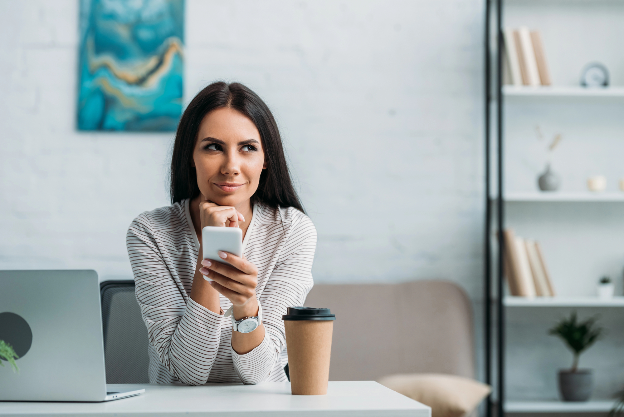 A woman with long dark hair sits at a desk holding a smartphone and smiling thoughtfully. She is in a modern, well-lit room with a laptop in front of her, a coffee cup next to her, a bookshelf in the background, and a painting on the wall.