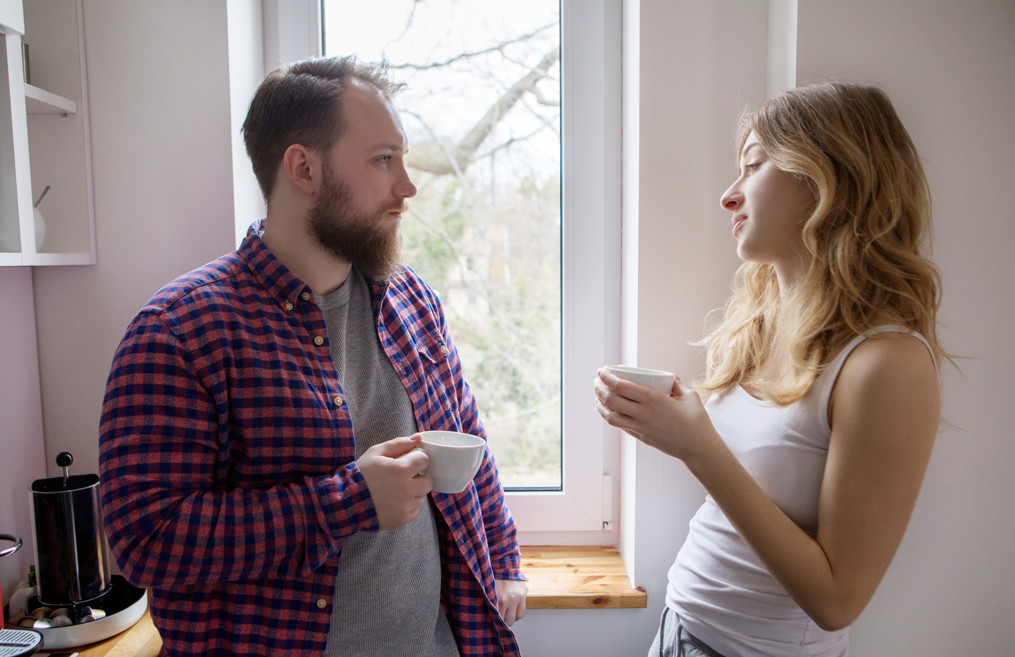 A man with a beard and a checked shirt, and a woman with long hair and a white tank top, are standing by a window in a bright room, holding white mugs and having a conversation.