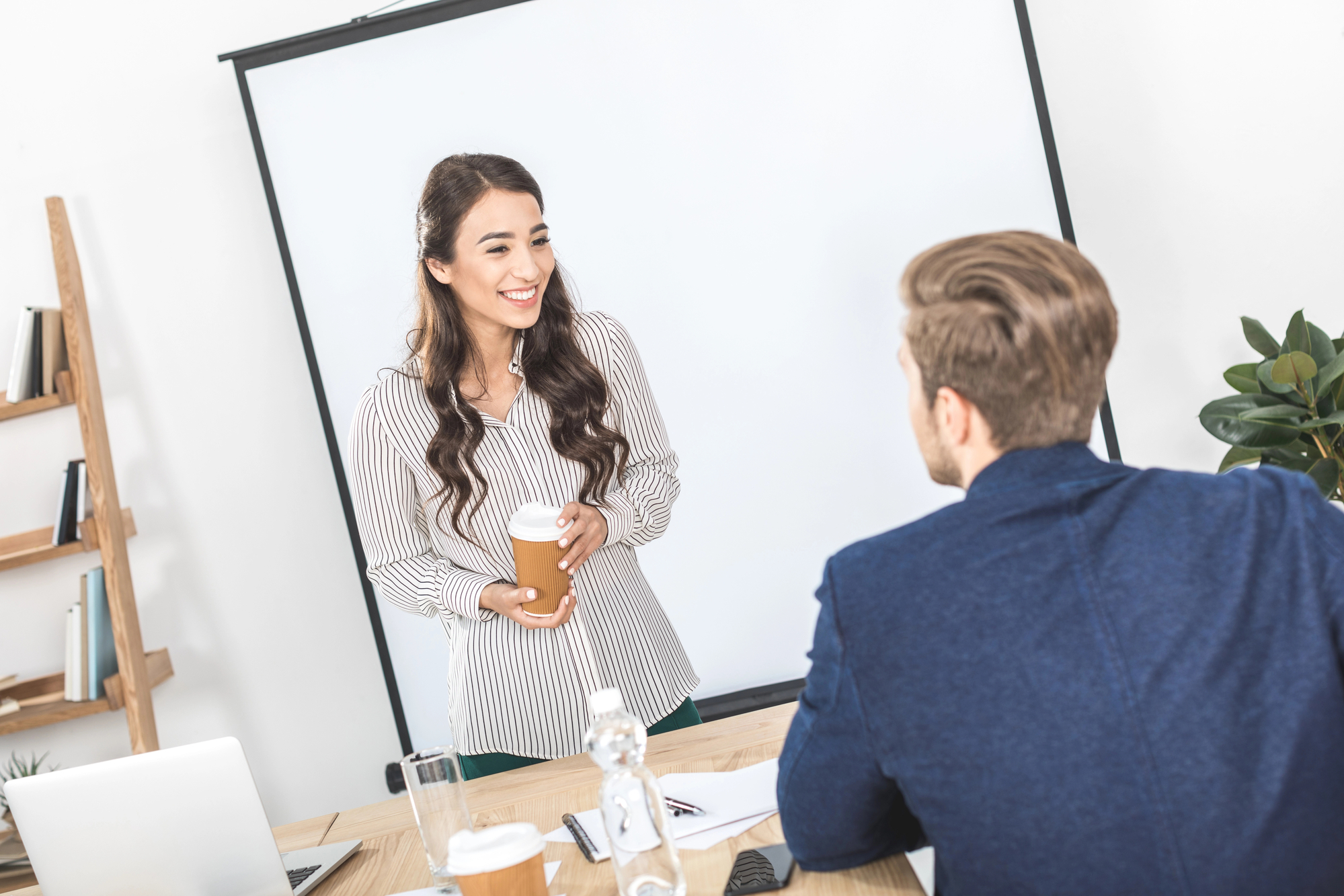 A woman holding a coffee cup smiles and talks to a man in a blue blazer. They are in a well-lit office with a projection screen in the background, a wooden shelf with books, and a desk with papers and a laptop. A plant is visible on the right.