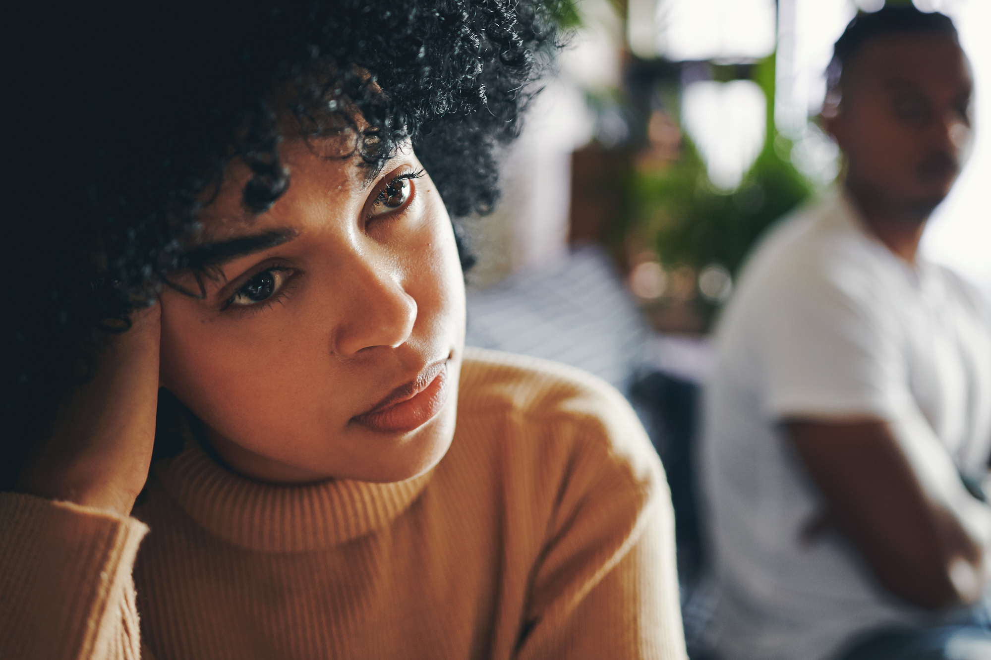 A woman with curly hair and a pensive expression rests her head on her hand, looking into the distance. In the blurred background, a man sits with his arms crossed, facing away. The scene suggests a moment of contemplation or emotional tension.