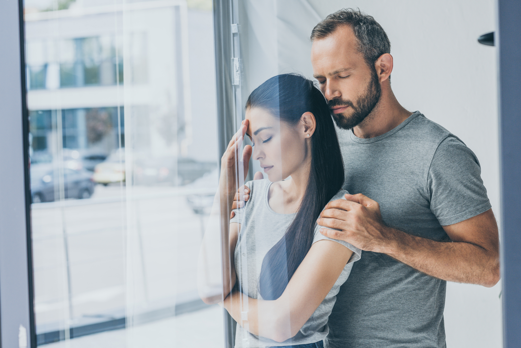 A man and woman stand together in a room near a window. The woman appears distressed with her hand on her forehead, while the man gently places his hands on her shoulders, offering comfort. Both are wearing casual grey t-shirts, with a blurred cityscape outside.