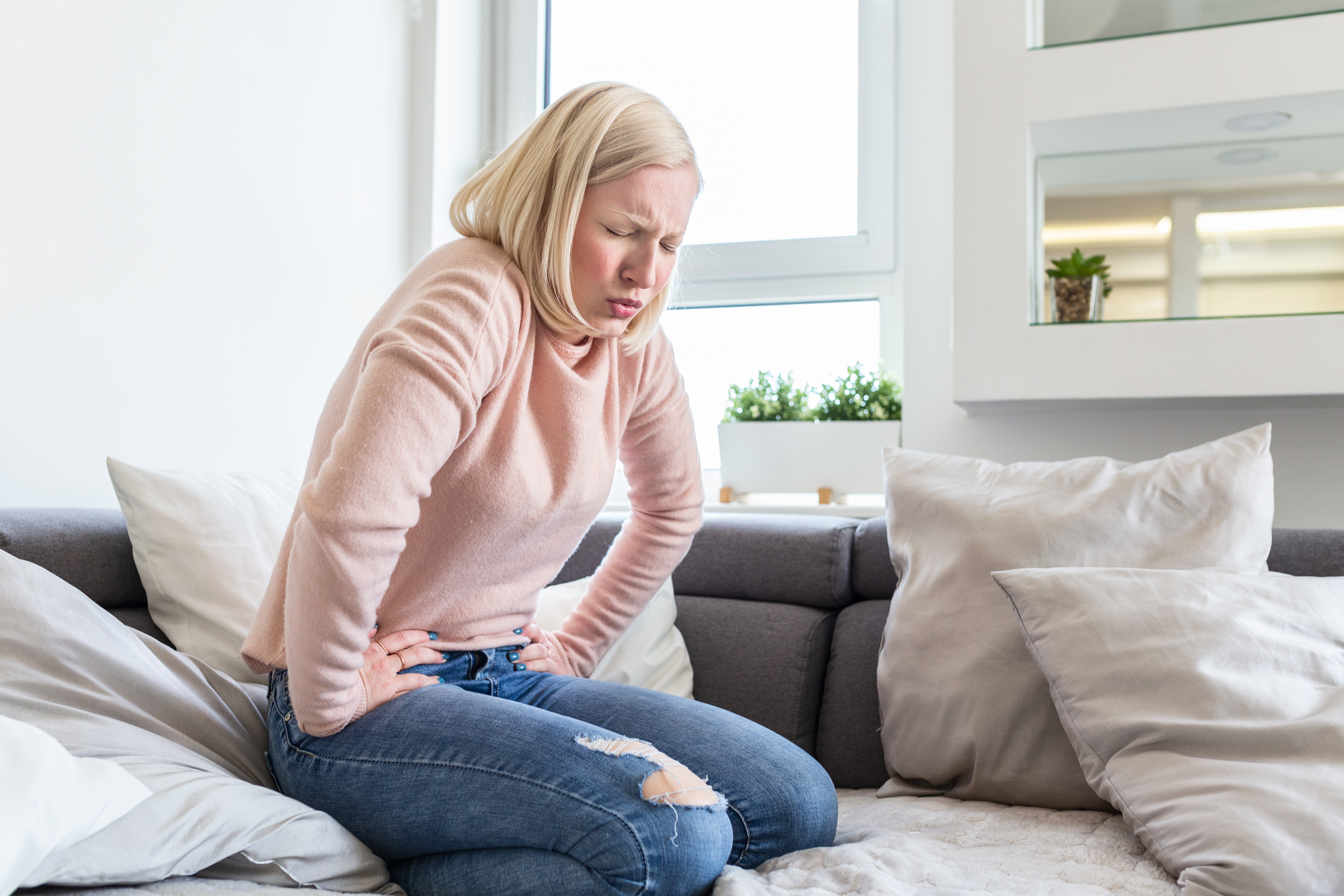 A woman with blonde hair wearing a pink sweater and blue jeans sits on a gray couch with several white and gray pillows, holding her stomach and appearing to be in pain.