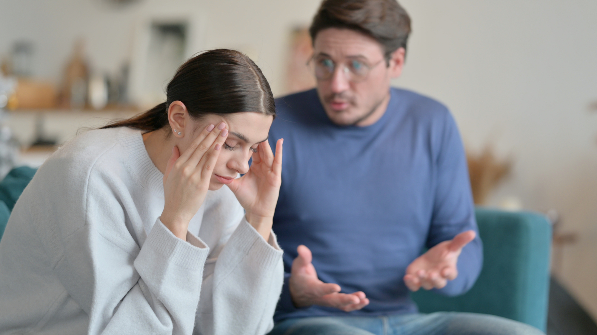 A woman sits on a couch, looking down and holding her temples with her fingers, appearing stressed. Next to her, a man in glasses and a blue sweater gestures with his hands and speaks, looking concerned. They are in a well-lit room with blurred background.