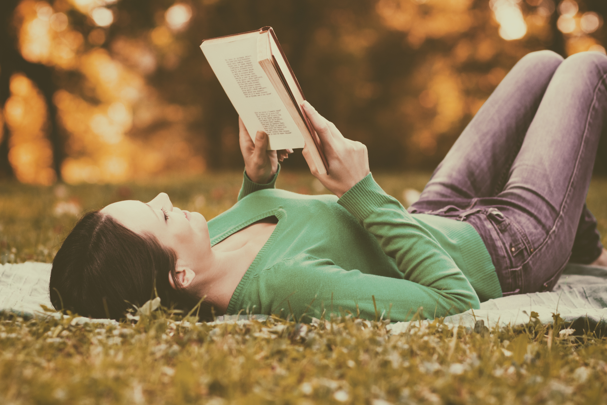 A woman in a green sweater and jeans is lying on a blanket in the grass, reading a book. The background is filled with blurred autumn foliage.