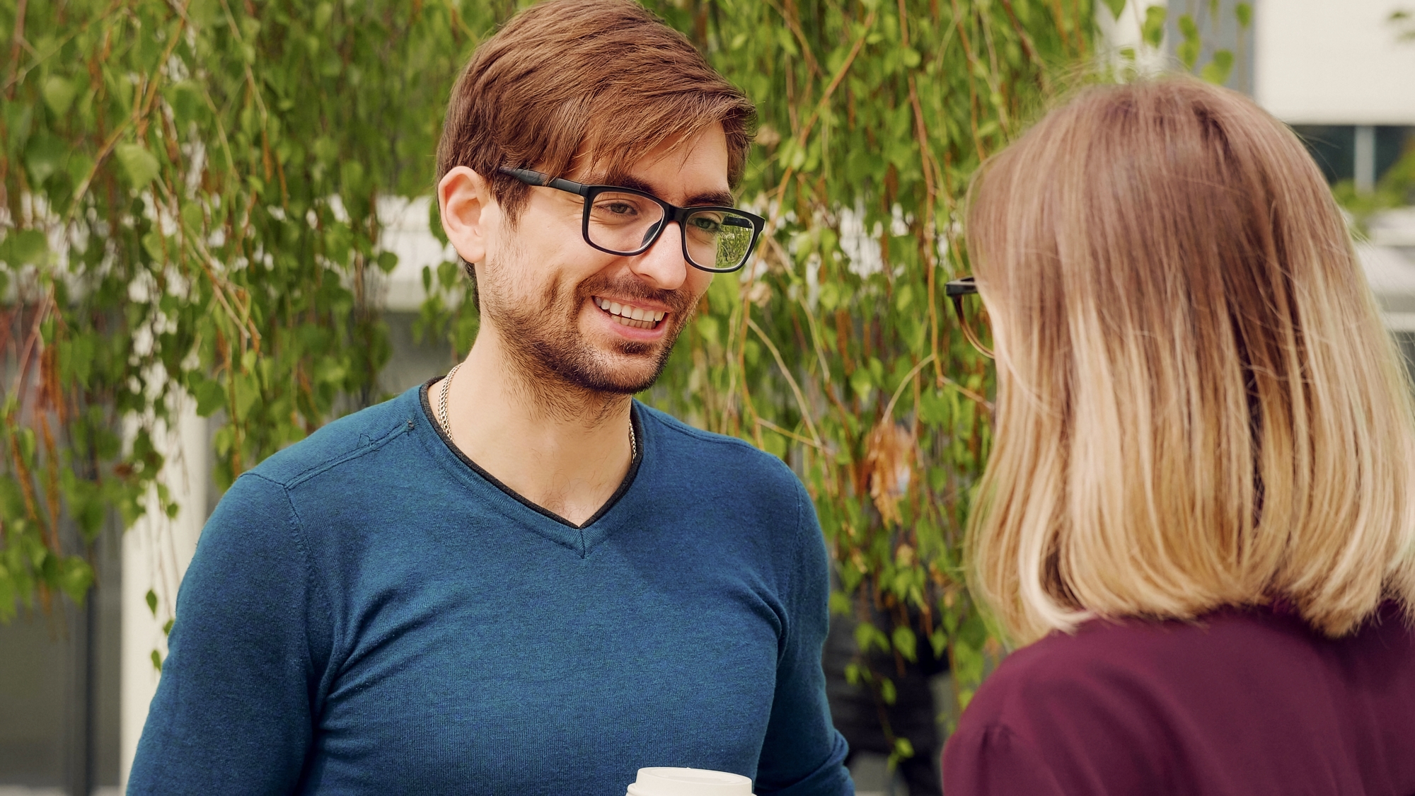 A man in a blue shirt and glasses smiles while talking to a woman with shoulder-length blonde hair and glasses. They are standing outdoors near some greenery. The man is holding a white cup.